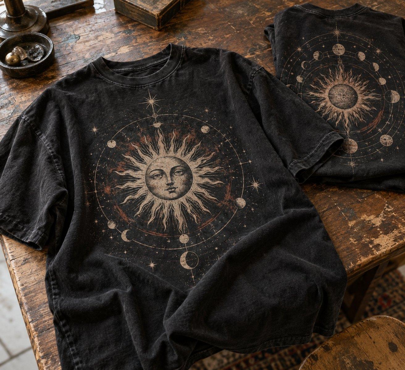 Washed black tee with a celestial sun and moon-phase print, laid on a wooden table beside its folded back